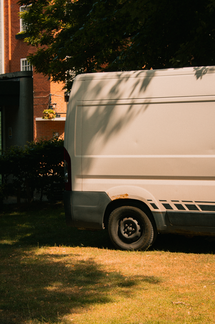 A bright orange vintage van parked on a residential street in front of a light-colored building with bay windows and a wooden door. The van has tinted side windows, black side mirrors, and a white roo