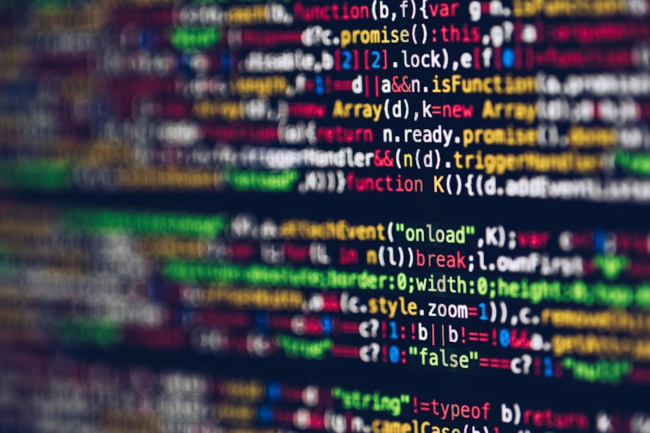 Close-up of a person's hand wearing a wristwatch, typing on a laptop keyboard with a black screen displaying lines of code or HTML. The laptop is positioned on a wooden surface, which appears to be a