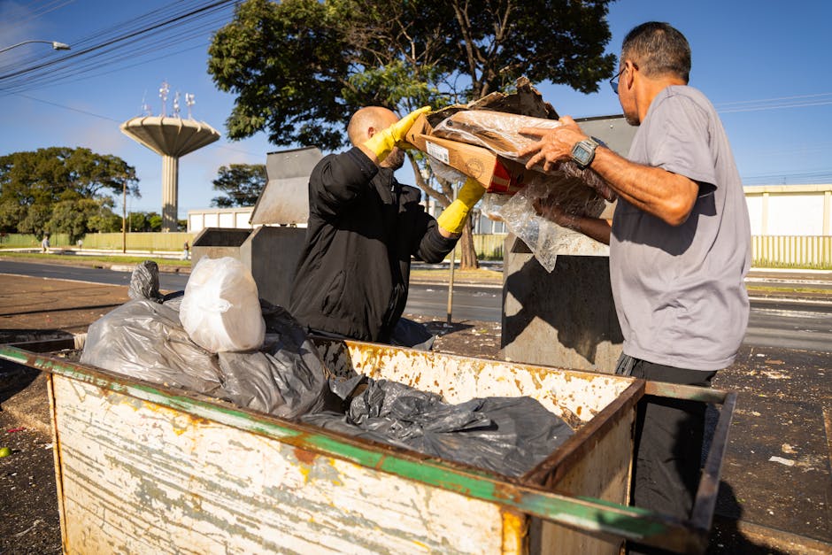 Two men involved in house removal or relocation work are standing outdoors beside a large, rusty metal dumpster filled with black and grey plastic trash bags and other waste materials. One man, wearin