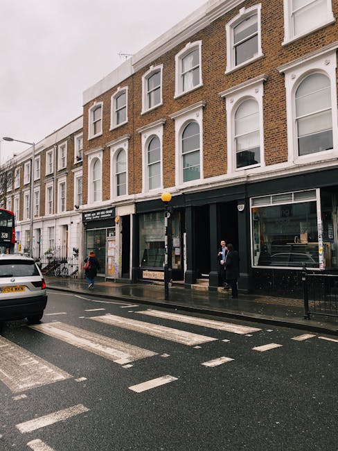 A street scene in a residential area with three-storey terraced brick buildings, some with white decorative window frames, on a cloudy day. In front of the buildings, there is a black pavement with a